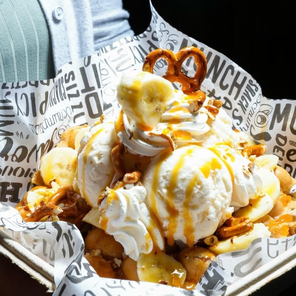 A large dessert with ice cream, whipped cream, bananas, pretzels, and caramel drizzle in a paper tray.