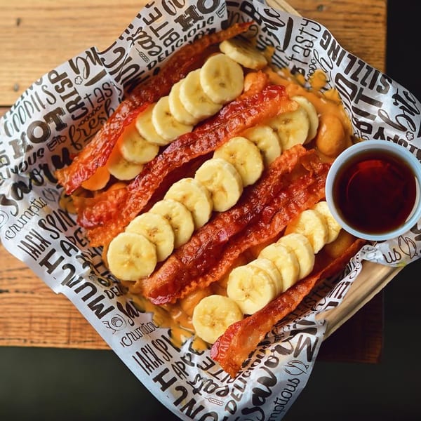 Crispy bacon strips and banana slices on a waffle, with a side of syrup.