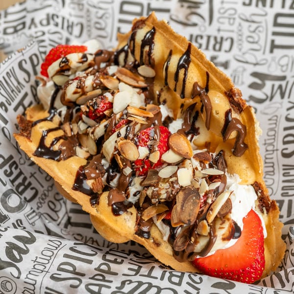 Bubble waffle with strawberries, bananas, whipped cream, chocolate drizzle, and almonds in a paper cone.