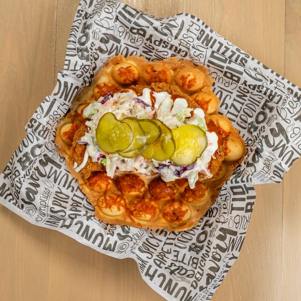 Crispy fried chicken, coleslaw, and pickles on a bubble waffle, served on patterned paper.