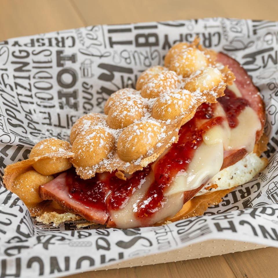 Bubble waffle sandwich with ham, melted cheese, and raspberry jam, dusted with powdered sugar.